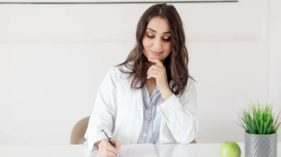 A female nutritionist in a lab coat writing notes with an apple and plant on the desk.